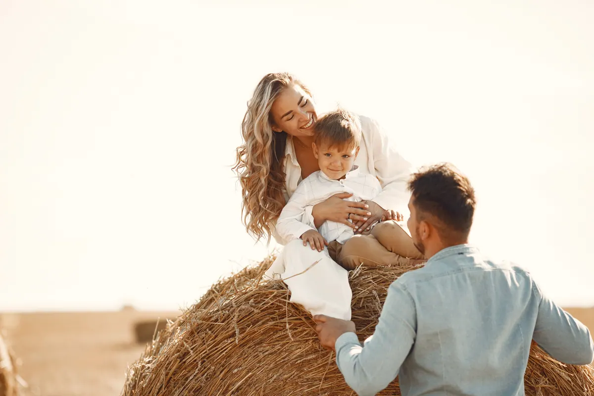 A mother and her small son sitting on a round hay bale while the father stands beside them