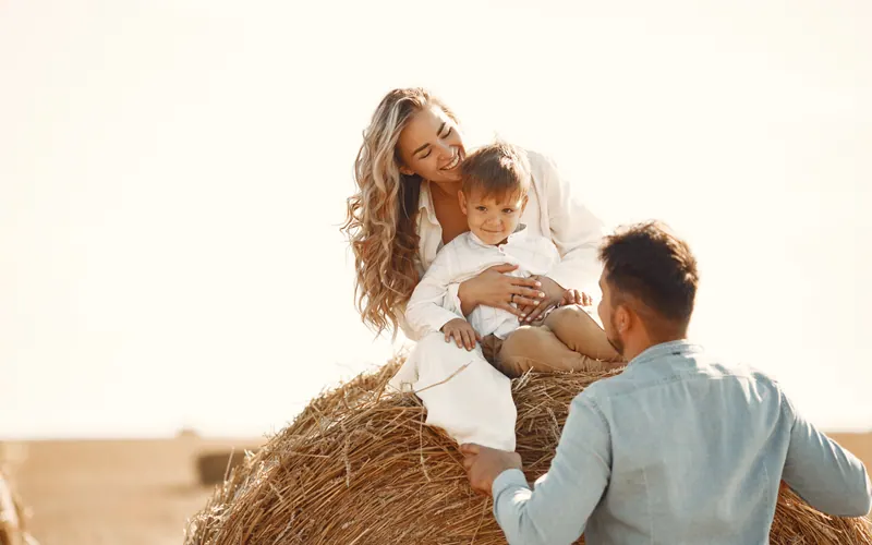 A mother and her small son sitting on a round hay bale while the father stands beside them