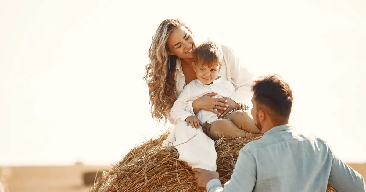 A mother and her small son sitting on a round hay bale while the father stands beside them