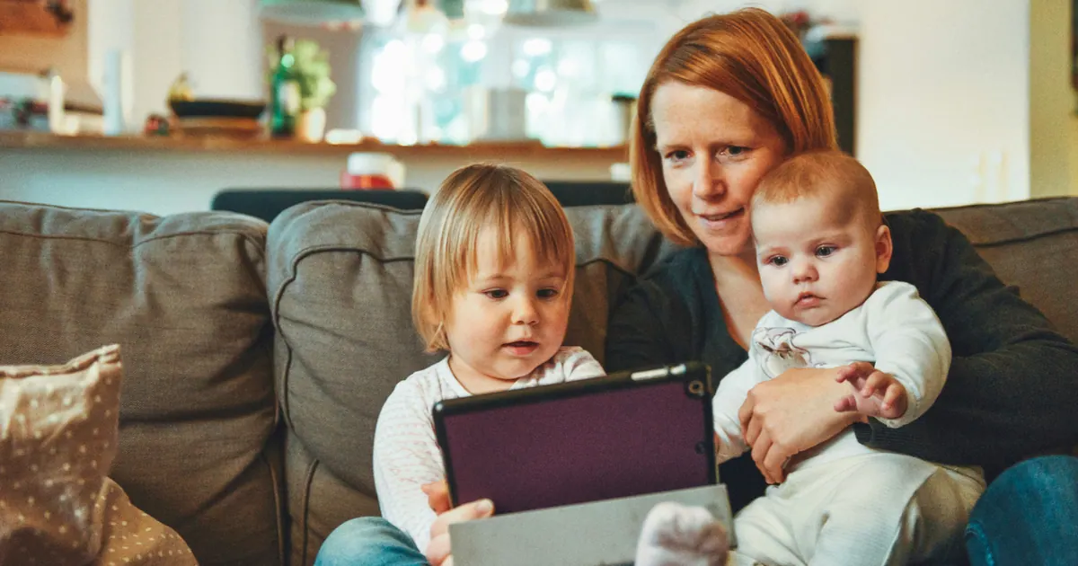 A mother sitting on a sofa with a toddler and a baby, holding a tablet