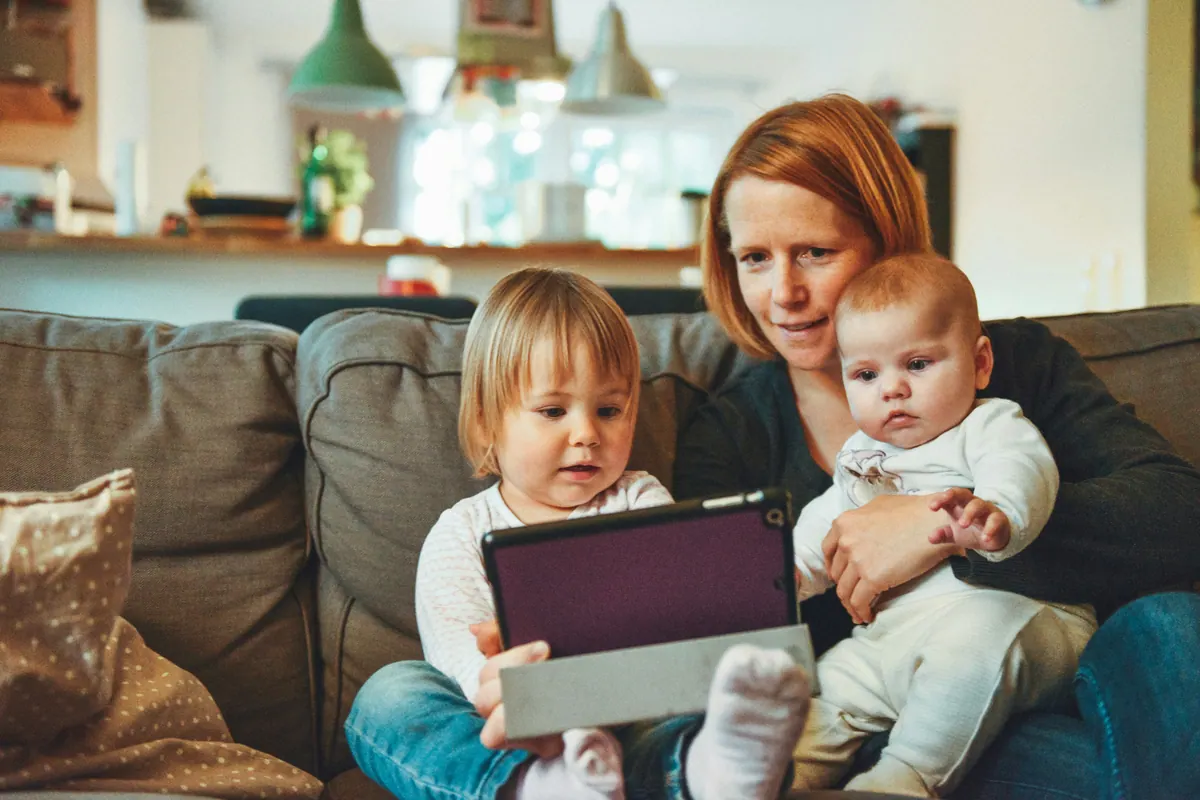 A mother sitting on a sofa with a toddler and a baby, holding a tablet