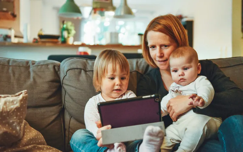 A mother sitting on a sofa with a toddler and a baby, holding a tablet