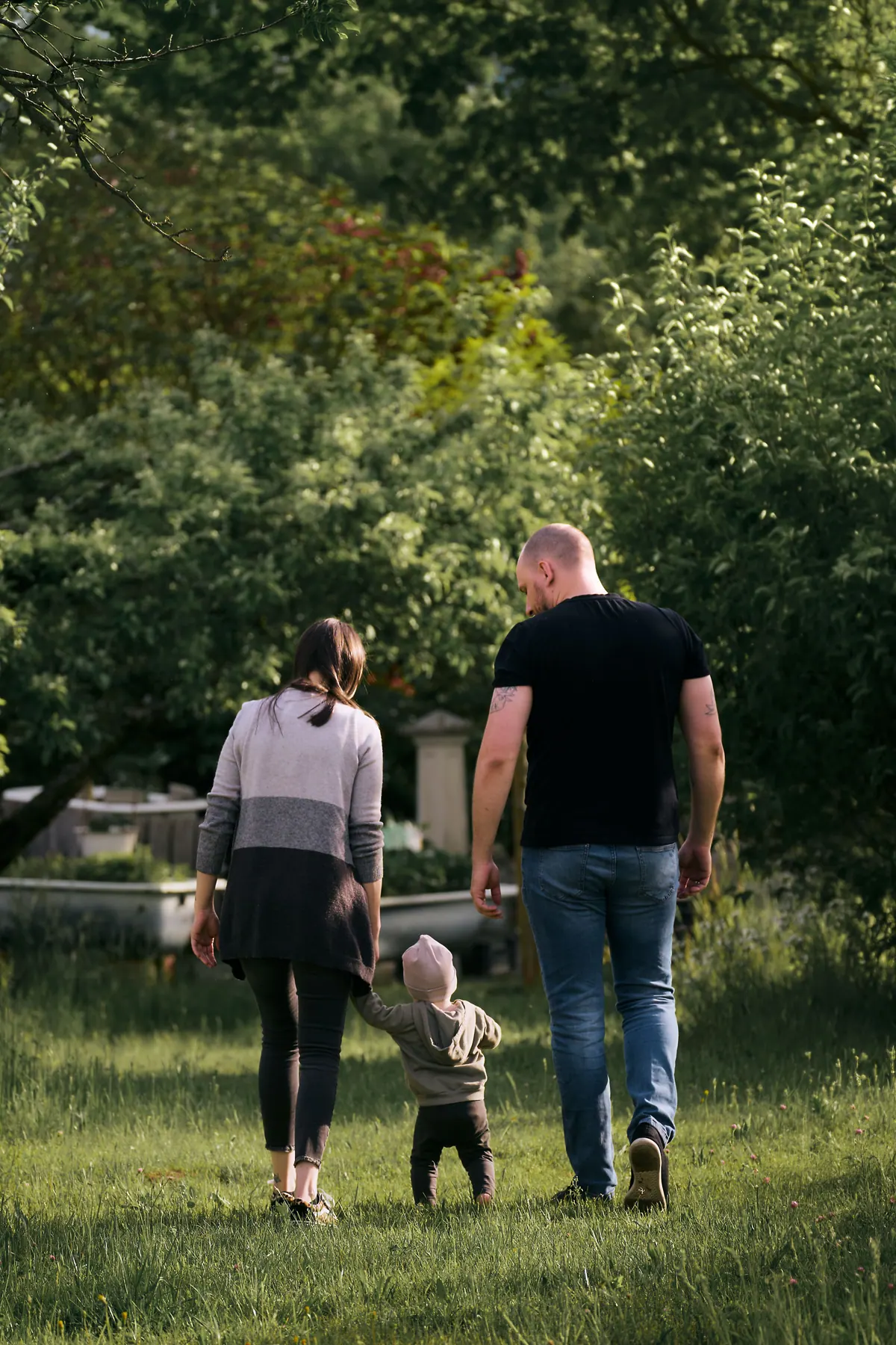 Two parents and a toddler walking away across a green garden, seen from behind