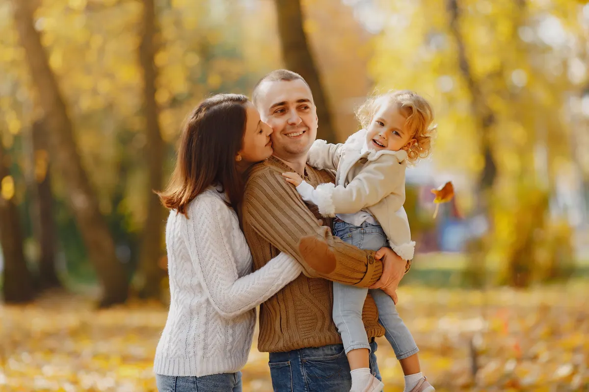 A mother kissing a father's cheek as he holds their toddler in an autumn park