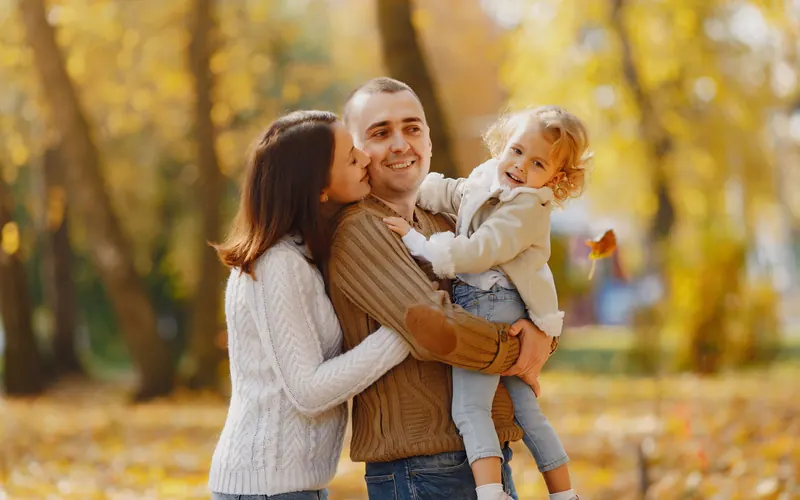 A mother kissing a father's cheek as he holds their toddler in an autumn park