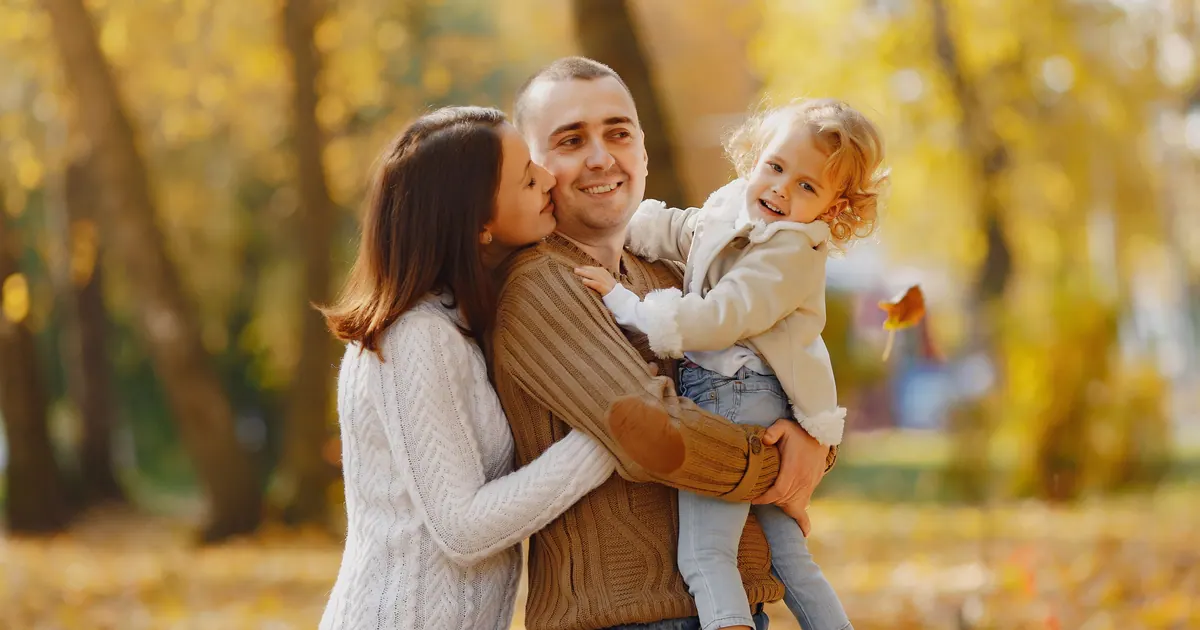 A mother kissing a father's cheek as he holds their toddler in an autumn park