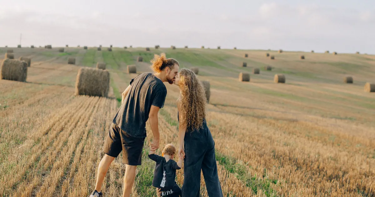 Two parents leaning together over a small child in a harvested field of hay bales