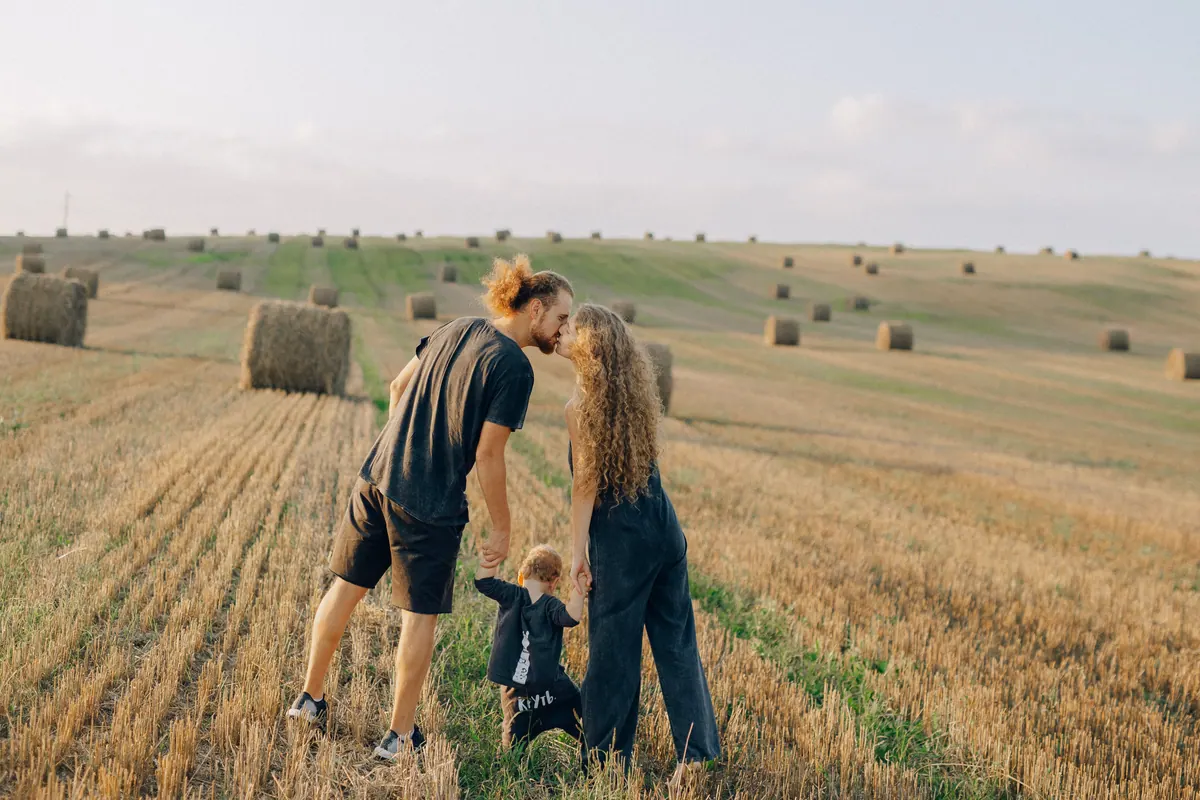 Two parents leaning together over a small child in a harvested field of hay bales