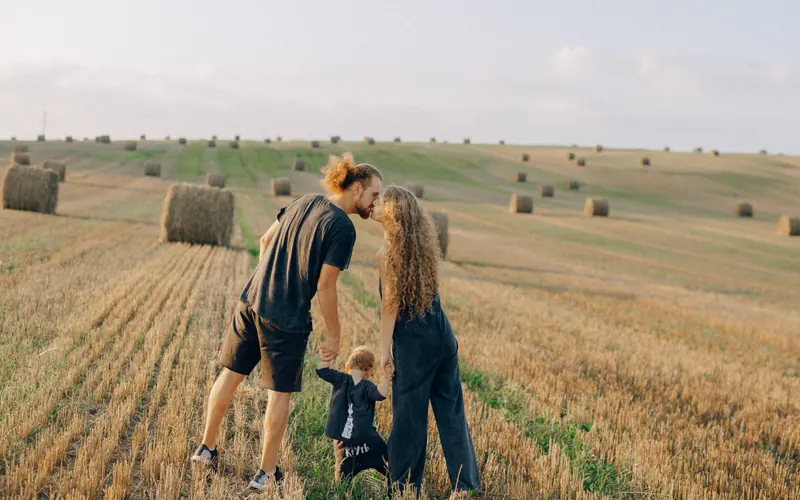 Two parents leaning together over a small child in a harvested field of hay bales