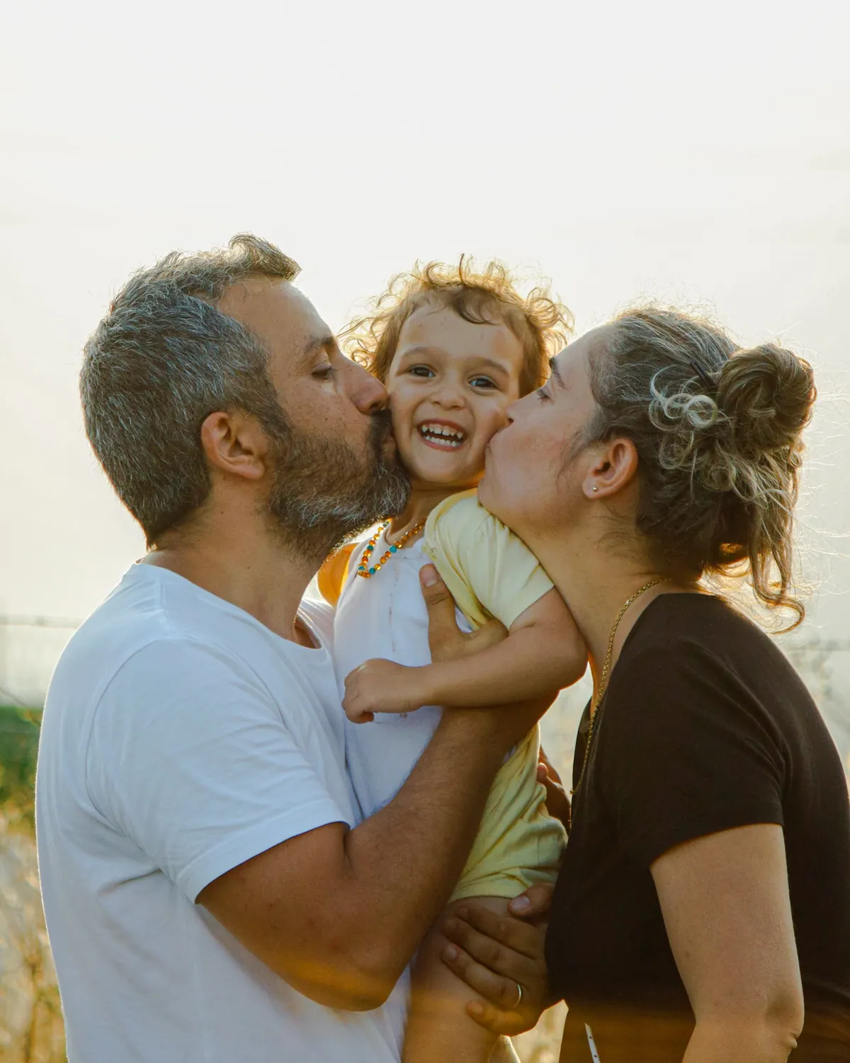 Two parents kissing the cheeks of a laughing toddler held between them in low sunlight