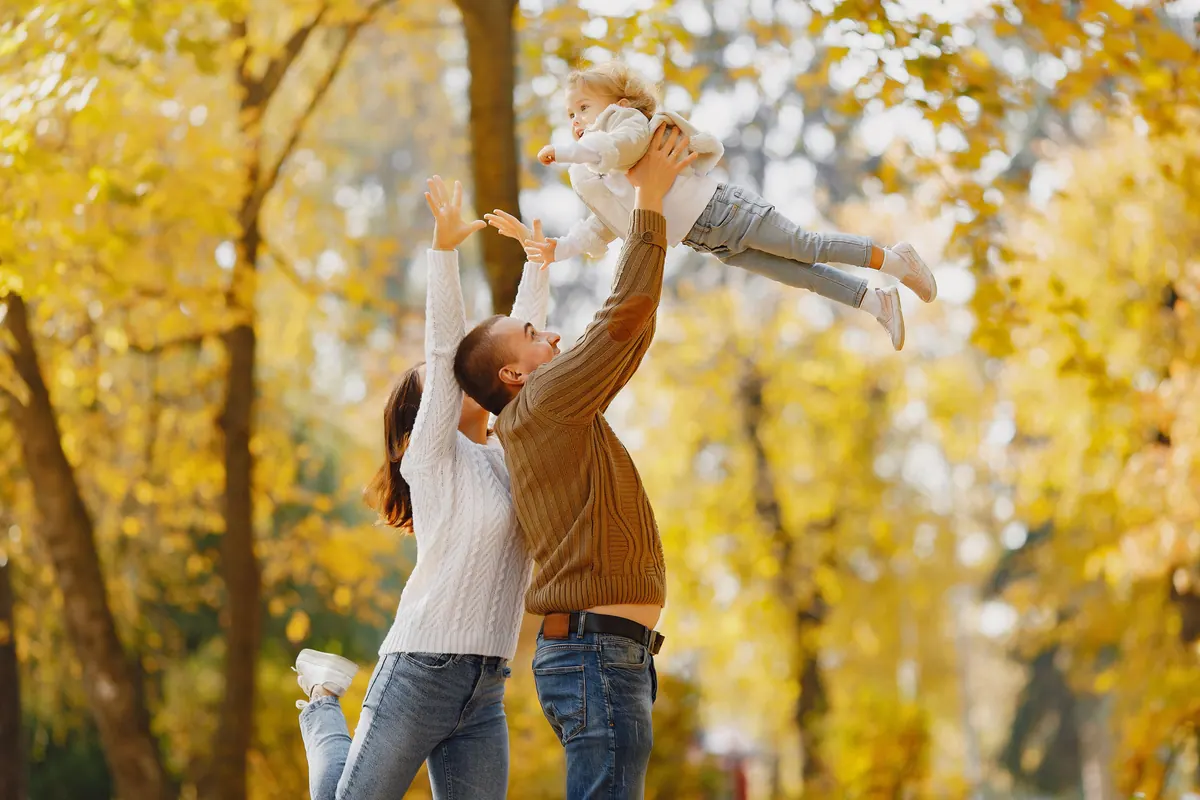 Two parents lifting a baby into the air beneath bright yellow autumn trees