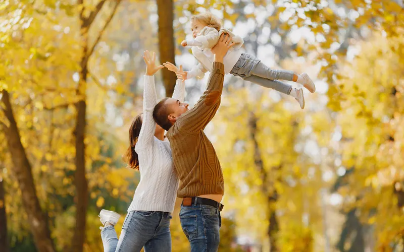 Two parents lifting a baby into the air beneath bright yellow autumn trees