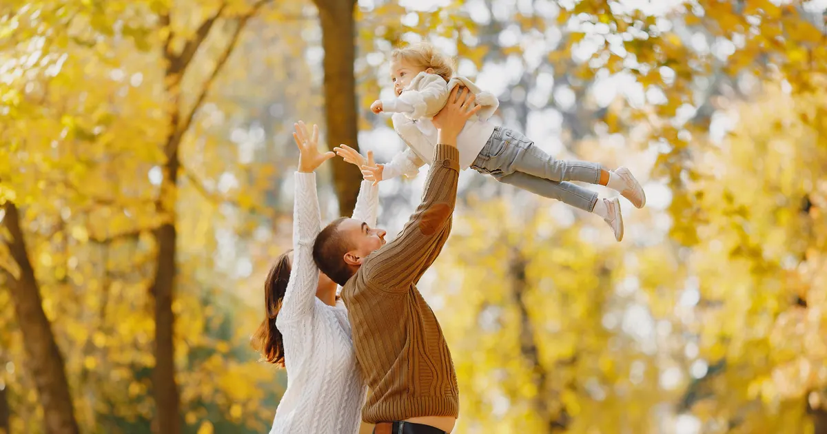 Two parents lifting a baby into the air beneath bright yellow autumn trees