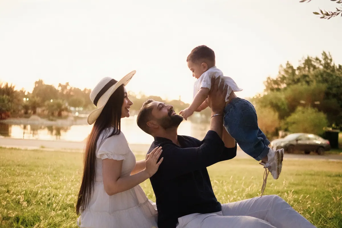 A father lifting a toddler overhead while the mother watches, in a park at golden hour