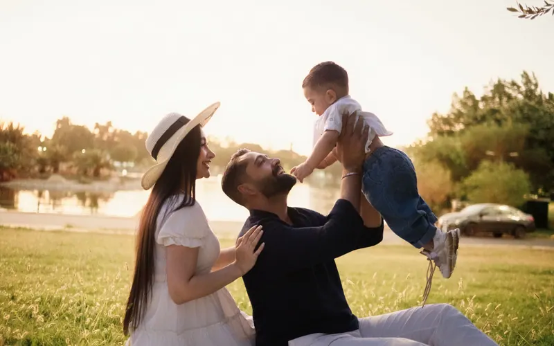 A father lifting a toddler overhead while the mother watches, in a park at golden hour