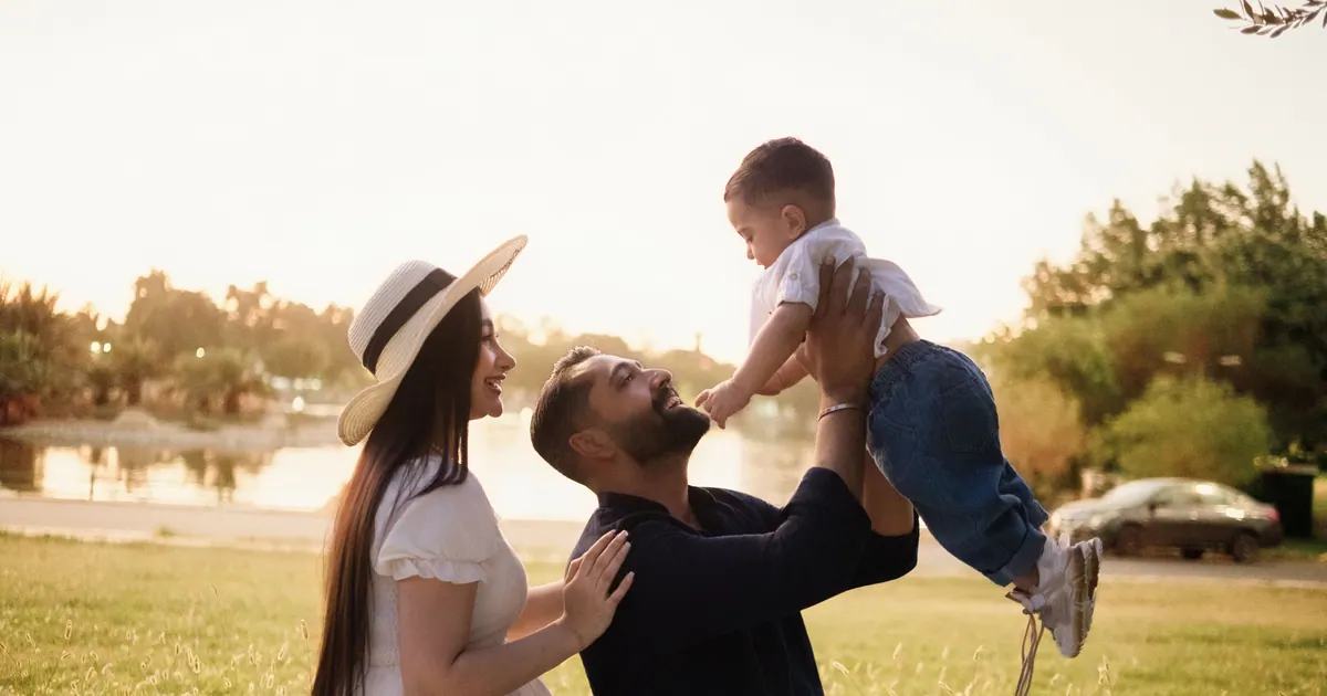 A father lifting a toddler overhead while the mother watches, in a park at golden hour