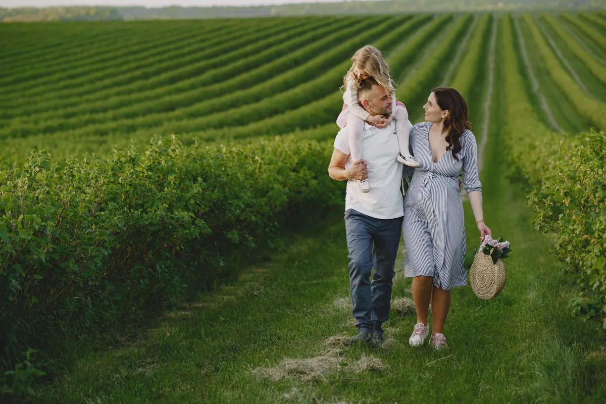Two parents walking a grass path between fields with a small child on one of their shoulders