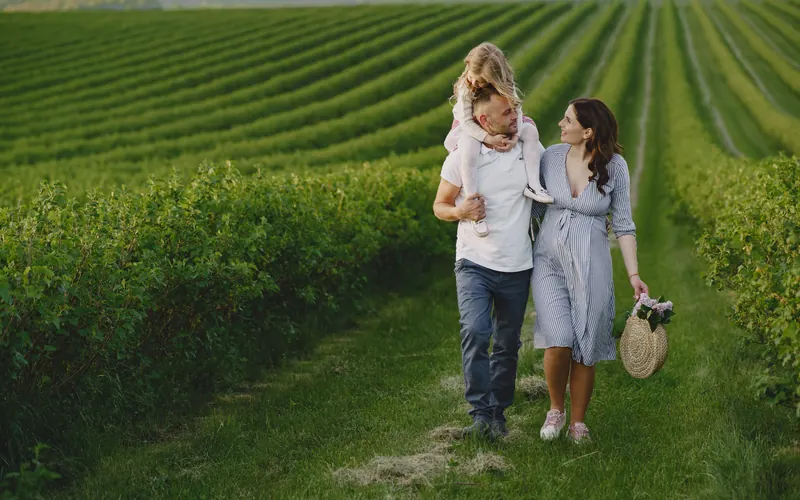 Two parents walking a grass path between fields with a small child on one of their shoulders