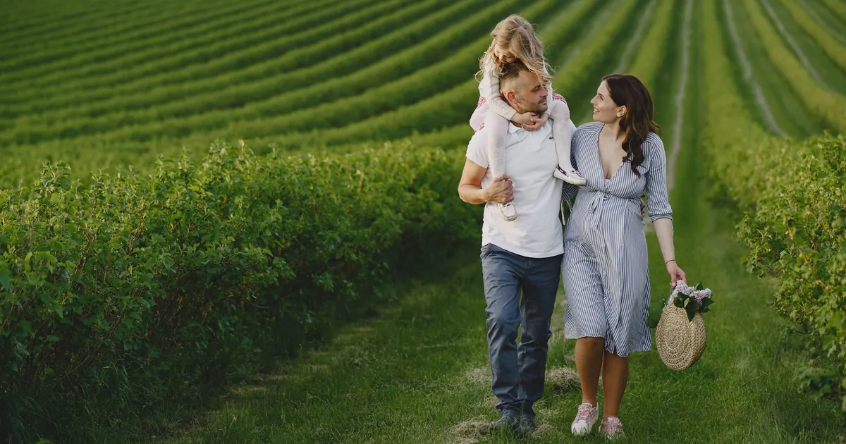 Two parents walking a grass path between fields with a small child on one of their shoulders