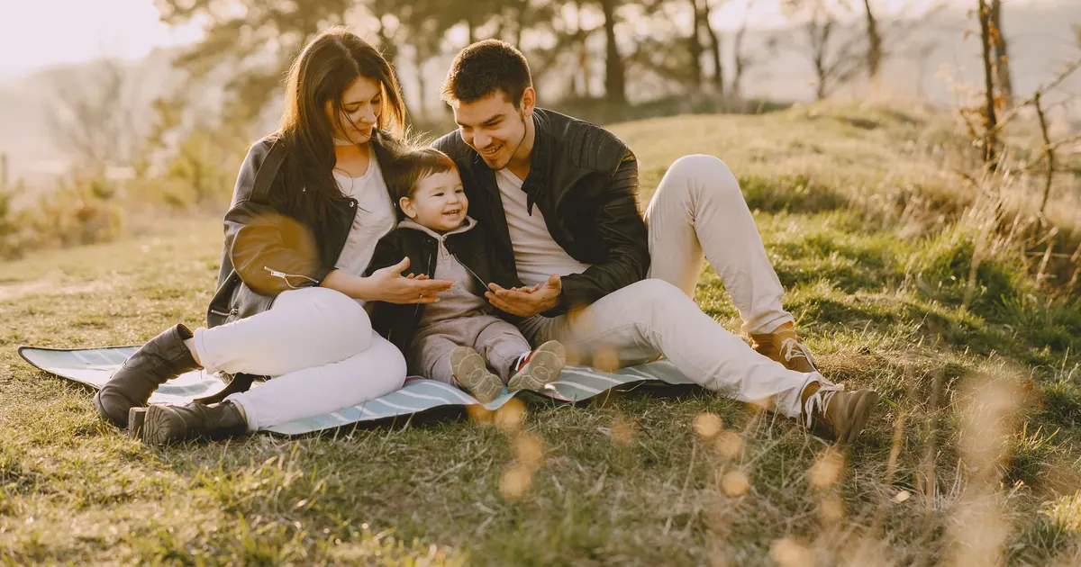Two parents sitting on a blanket on a hillside with their baby between them