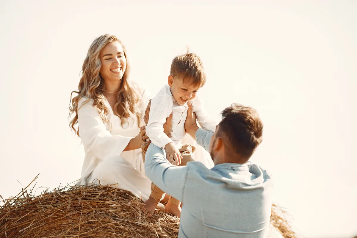 Two parents playing with their toddler on a round hay bale in a harvested field