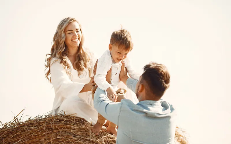 Two parents playing with their toddler on a round hay bale in a harvested field