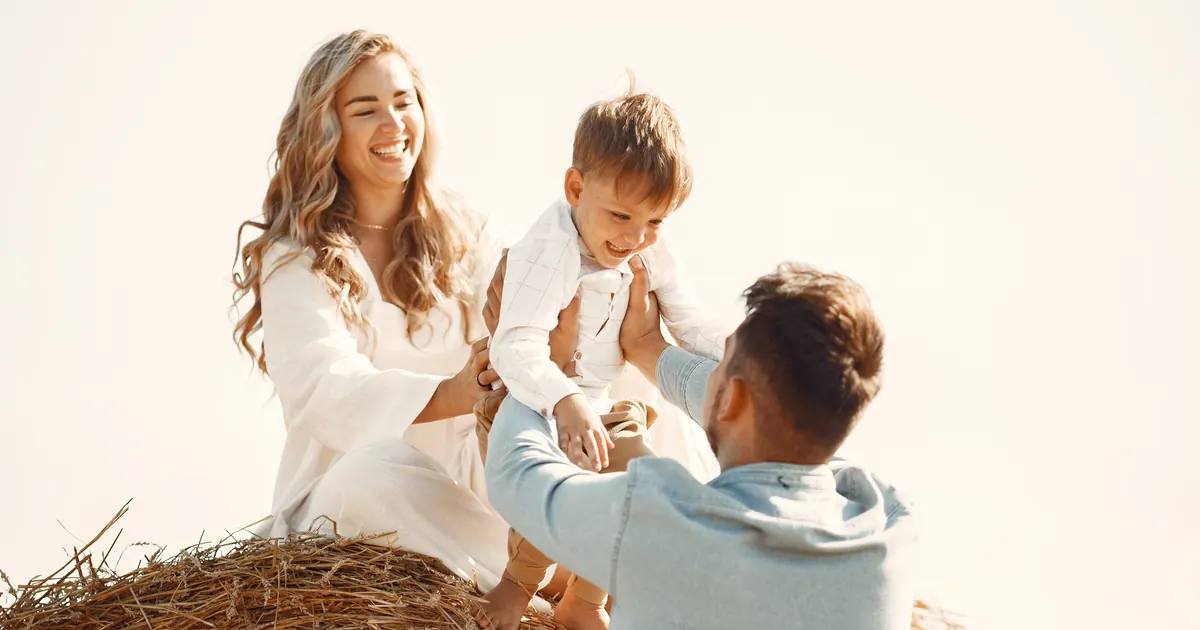 Two parents playing with their toddler on a round hay bale in a harvested field