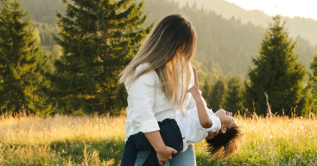 A woman carrying a laughing child on her back in a sunlit meadow