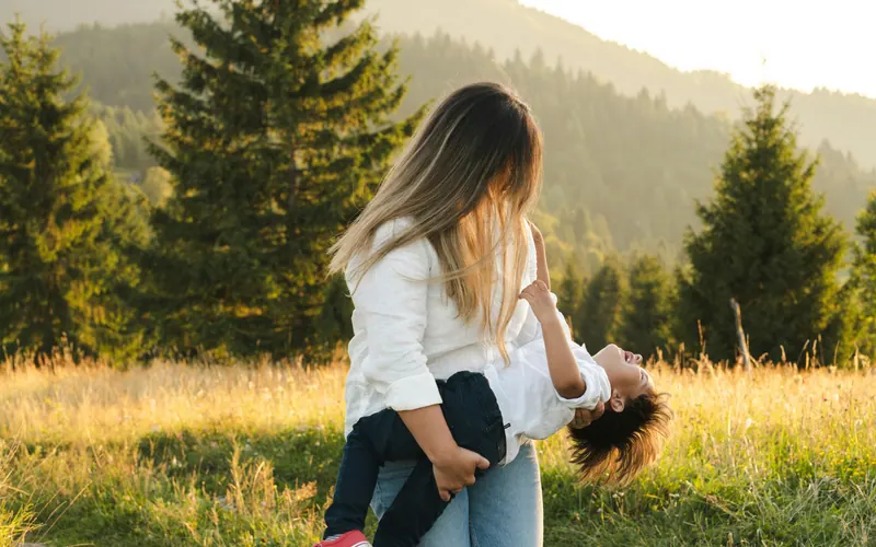 A woman carrying a laughing child on her back in a sunlit meadow